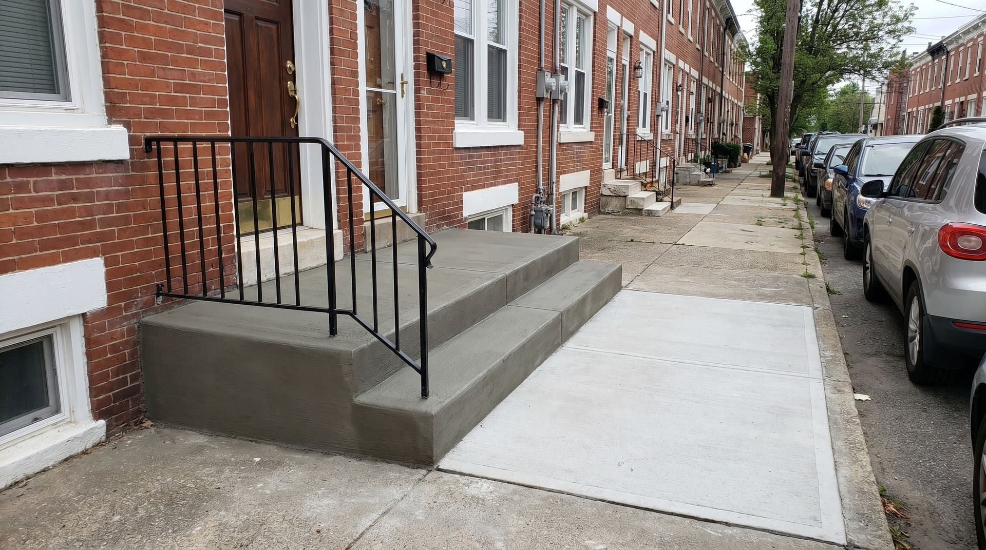 Freshly rebuilt concrete front stoop with handrail and adjacent new sidewalk panel on a 1910s red-brick rowhouse on a dense Bridgeport Pennsylvania residential street