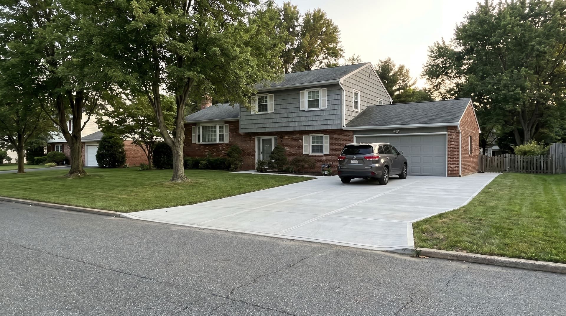 1972-era King of Prussia split-level home with red brick lower level and gray clapboard upper, with a brand-new wide concrete driveway running to the attached garage in late afternoon natural light