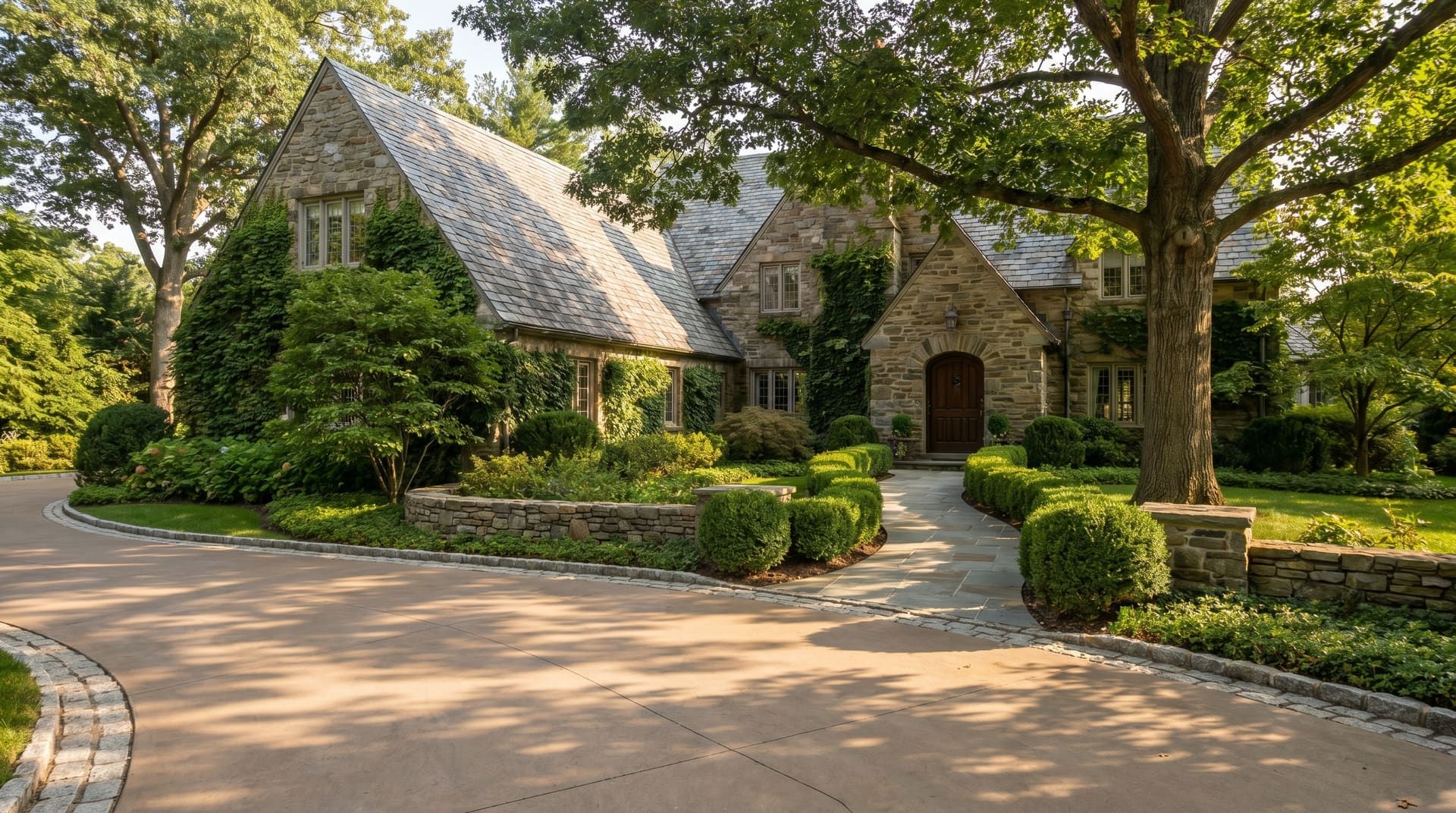 1920s stone Tudor Revival estate home on a third-acre Lower Merion Main Line lot with mature trees and ivy, a long curved decorative concrete driveway with stamped Belgian-block border, and a Pennsylvania bluestone front walk