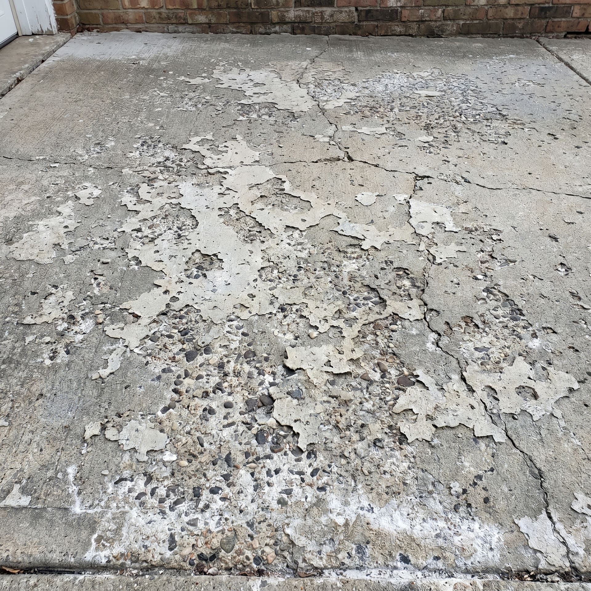 Failed concrete driveway with surface scaling and spalling across a wide section, top layer flaking off in irregular patches exposing darker aggregate underneath, salt damage residue visible