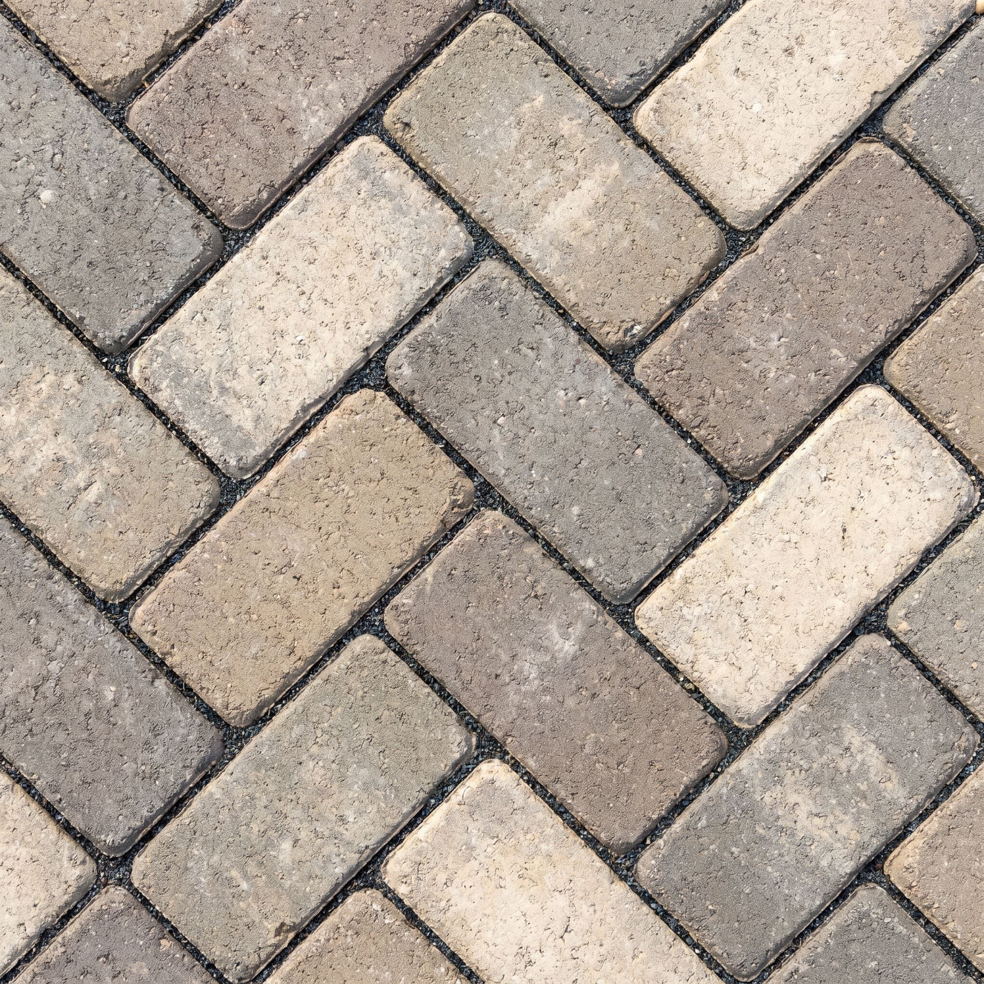 Close-up detail of concrete pavers laid in a tight herringbone pattern in muted natural-stone tones with tight polymeric joint sand in the seams