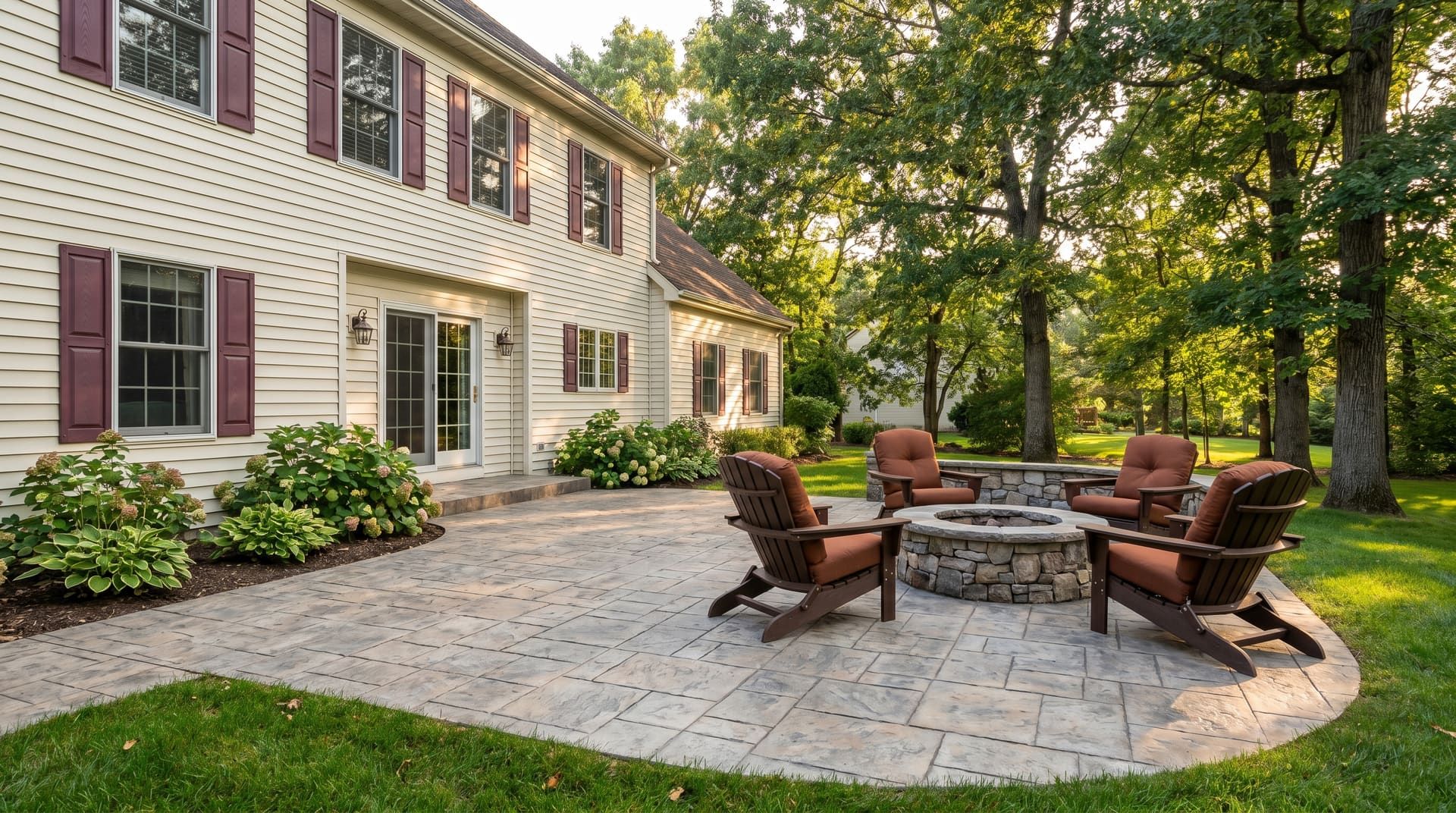Stamped concrete patio in an ashlar-slate pattern behind a 1990s King of Prussia colonial home with cream siding and burgundy shutters, with Adirondack chairs around a built-in stone fire pit at late-summer afternoon golden hour